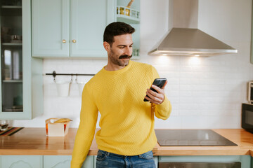 Handsome man in yellow sweater using mobile phone against kitchen counter