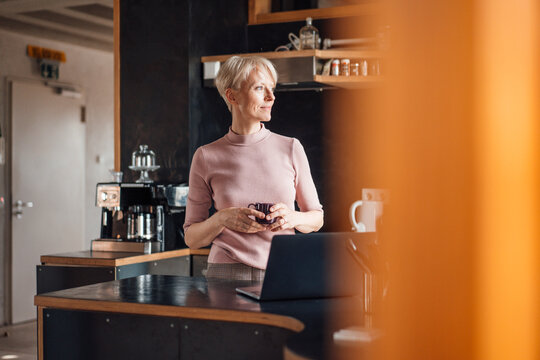 Smiling businesswoman looking away while holding coffee cup in kitchen at home office