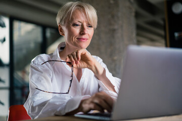 Smiling businesswoman holding eyeglasses while working on laptop at home office