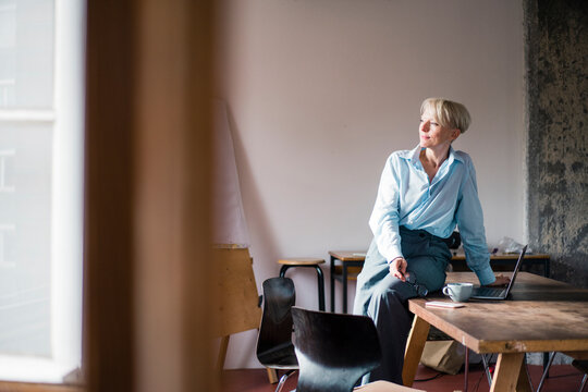 Smiling businesswoman with laptop sitting on desk while looking away at home office
