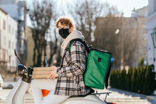 Food delivery service by scooter with courier concept. Curly young male in black protective mask with green thermal backpack waiting customer. Deliveryman holding pizza boxes with street on background