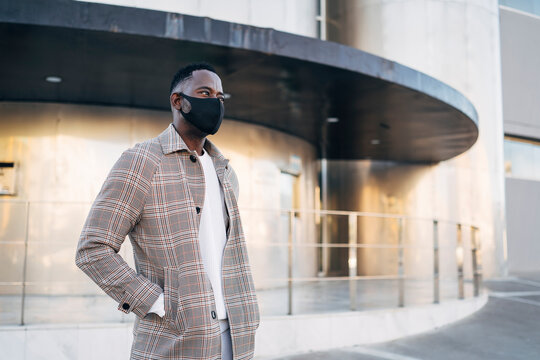 African man wearing protective face mask with hand in pocket standing against building