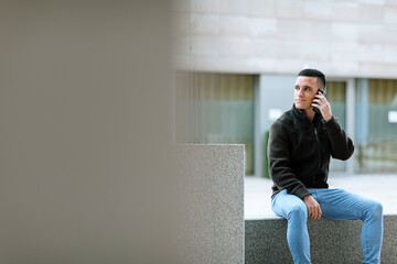 Young man talking on phone while sitting on bench against building