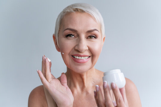 Smiling woman applying facial cream on face while standing against gray background