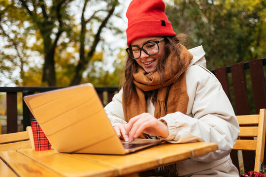 Woman wearing knit hat and scarf smiling while working on laptop at sidewalk cafe