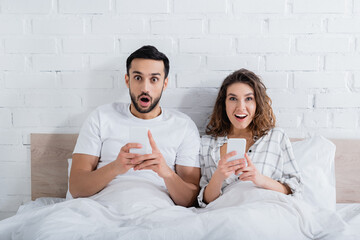 amazed interracial couple lying on bed and using smartphones.
