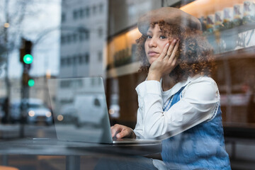Thoughtful teenage girl sitting with hand on chin by laptop at cafe window
