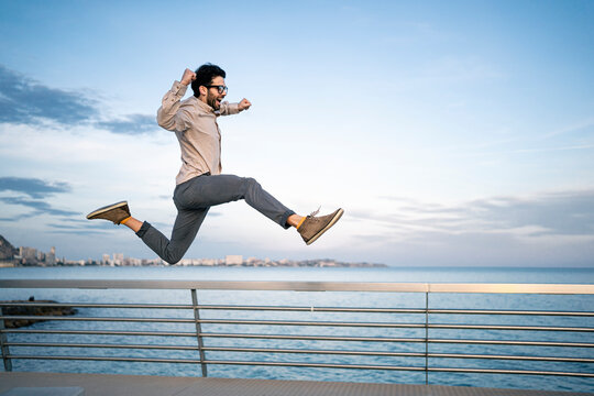 Carefree male entrepreneur with arms raised jumping by railing in front of blue sky
