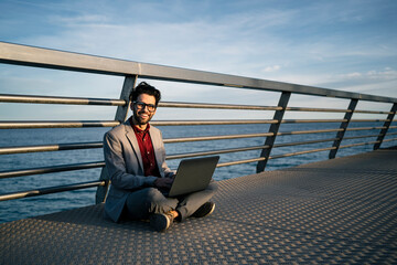 Smiling freelance worker with laptop sitting cross legged on pier