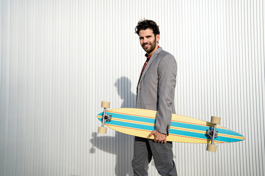 Smiling businessman with longboard standing by wall