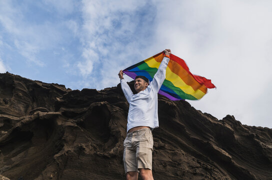 Smiling male tourist waving LGBTQI Flag while standing by rock at El golfo, Lanzarote, Spain
