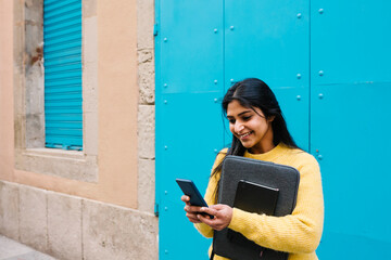 Smiling woman with digital tablet and laptop bag using mobile phone against blue wall