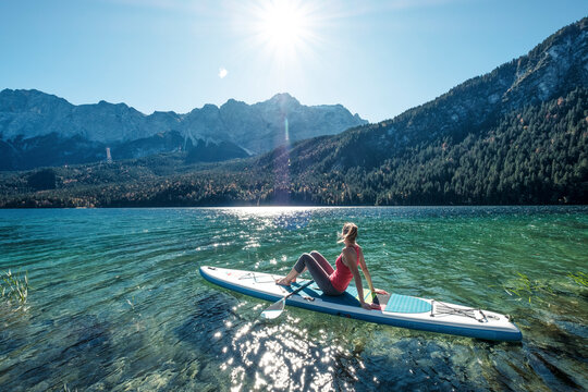 Germany, Bavaria, Garmisch Partenkirchen, Young woman sitting on stand up paddle board on Lake Eibsee and looking at Zugspitze Mountain