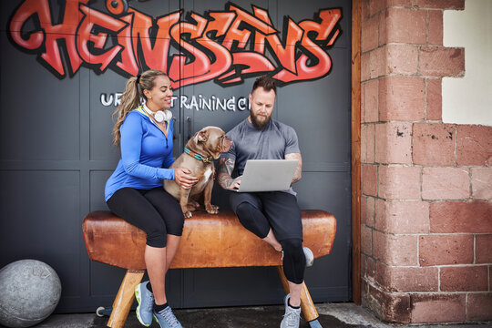 Male and female athlete using laptop while sitting with bulldog on bench against graffiti door