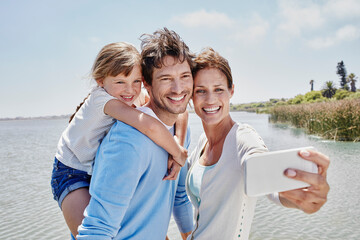 Smiling woman taking selfie with family by lake on sunny day
