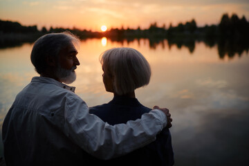 Romantic holiday. Senior loving couple sitting together on lake bank enjoying beautiful sunset.