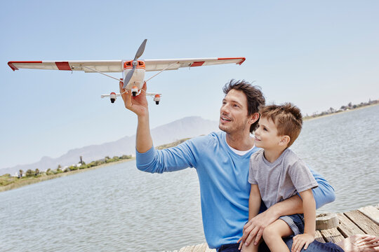 Father holding airplane toy while sitting with son by lake
