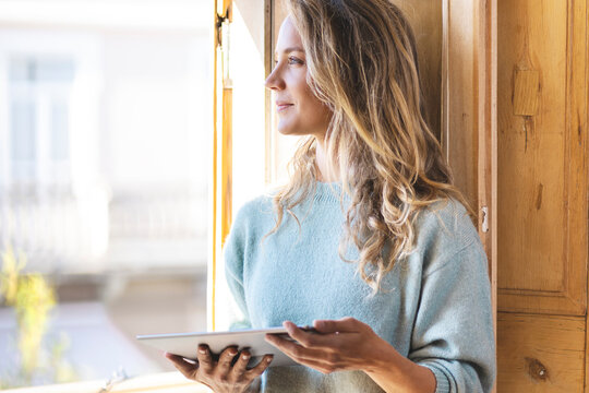 Smiling thoughtful woman with digital tablet looking out of window at home