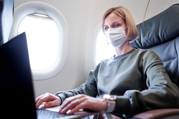 Young woman wearing face mask is traveling on airplane using laptop computer. New normal travel...