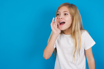 beautiful Caucasian little girl wearing white T-shirt over blue background look empty space holding hand near her face and screaming or calling someone.