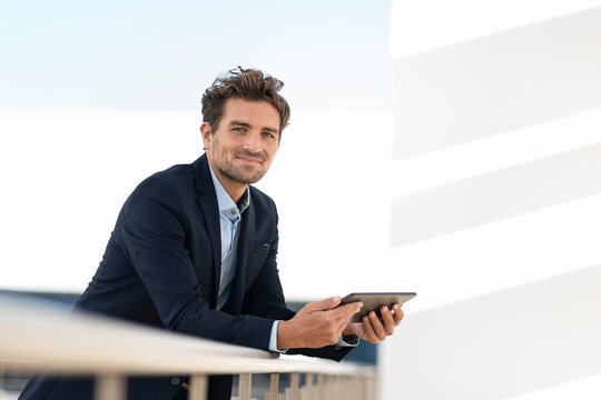 Smiling businessman holding digital tablet while standing by railing at office terrace