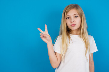 beautiful Caucasian little girl wearing white T-shirt over blue background makes peace gesture keeps lips folded shows v sign. Body language concept