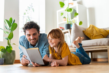 Smiling father and daughter reading book together while lying down on floor at home