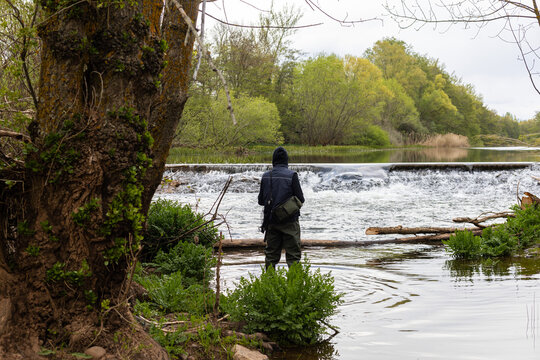 Young angler wearing a vest and high plastic boots, standing in the middle of the river to fish with a rod and tackle against the backdrop of a small waterfall in the photo