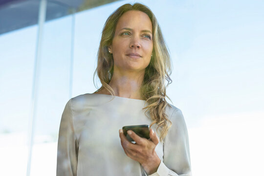 Female professional with mobile phone standing by glass window at office - Powered by Adobe