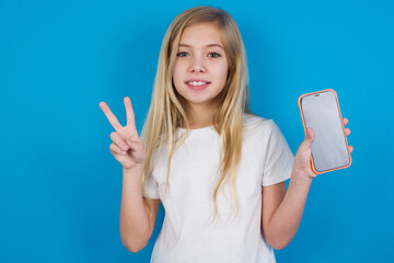 beautiful Caucasian little girl wearing white T-shirt over blue background holding modern device showing v-sign