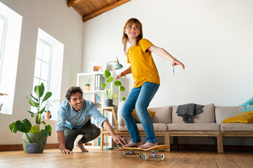 Father helping daughter to ride skateboard in living room at home