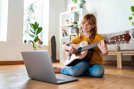 Cute redhead girl learning guitar with online tutorial through laptop in living room at home