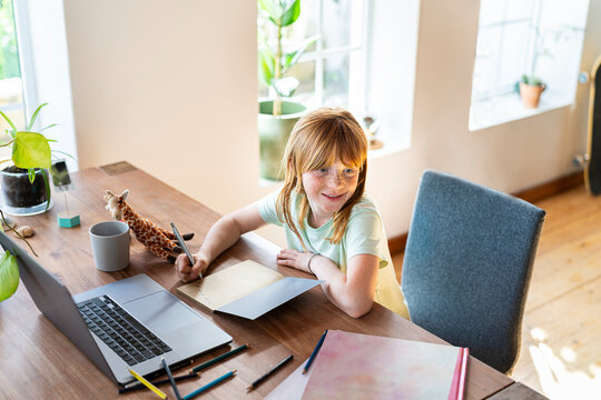 Smiling redhead girl looking away while doing homework in front of laptop at home
