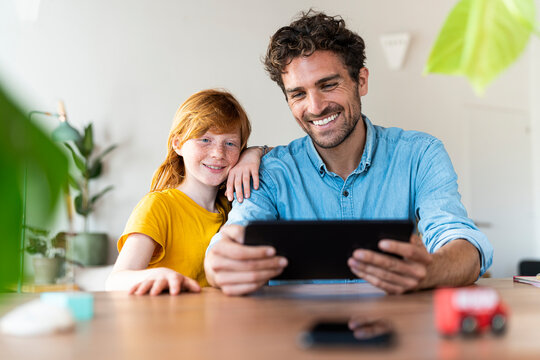Father with redhead daughter smiling and looking at digital tablet together in living room