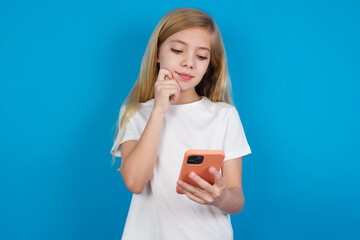Fototapeta premium Thoughtful happy beautiful Caucasian little girl wearing white T-shirt over blue background hold look phone, copyspace