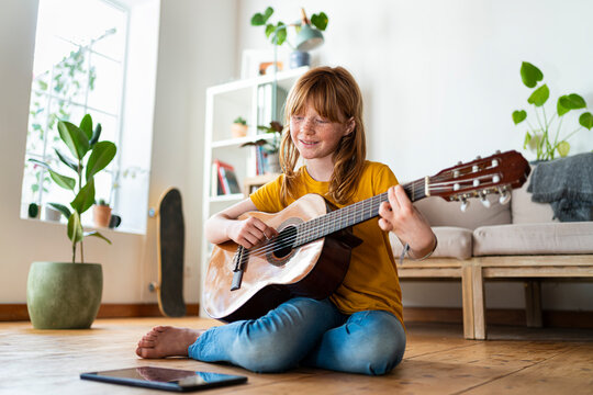 Redhead girl learning guitar online through digital tablet while sitting on floor at home