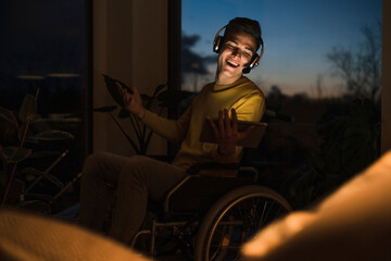 Happy ÔøΩoung man in wheelchair wearing wireless headphones while making video call through digital tablet in living room