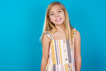 beautiful Caucasian little girl wearing stripped dress over blue background with broad smile, shows white teeth, feeling confident rejoices having day off.