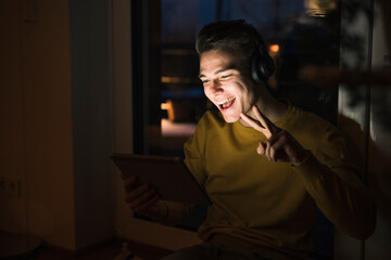 Cheerful man wearing headphones gesturing peace sign on video call while sitting in living room