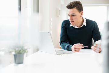 Young male professional with smart phone looking at laptop in home office