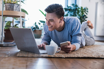 Smiling young with smart phone using laptop while lying on carpet in living room