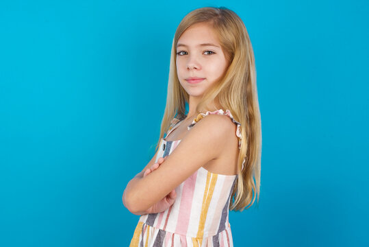 Image of cheerful beautiful Caucasian little girl wearing stripped dress over blue background with arms crossed. Looking and smiling at the camera. Confidence concept.