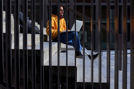 Disabled man with leg prosthesis using laptop while sitting on steps seen through metal railing