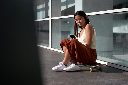 Asian woman using smart phone while sitting on skateboard during sunny day - Powered by Adobe