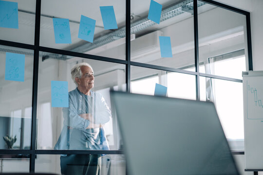 Senior entrepreneur with arms crossed reading note on glass wall while standing at office