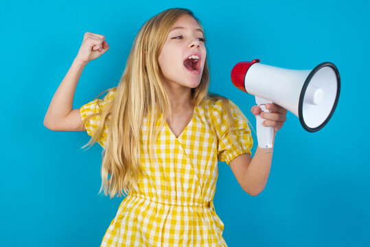 beautiful Caucasian little girl wearing yellow dress over blue background communicates shouting loud holding a megaphone, expressing success and positive concept, idea for marketing or sales.