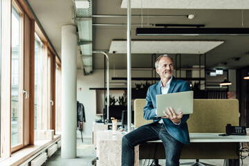 Businessman holding laptop while sitting on desk in office