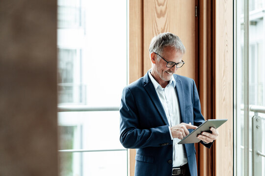 Smiling businessman using digital tablet while standing against glass window
