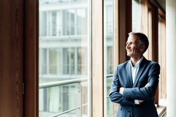 Contemplating businessman looking away while standing with arms crossed at window in office