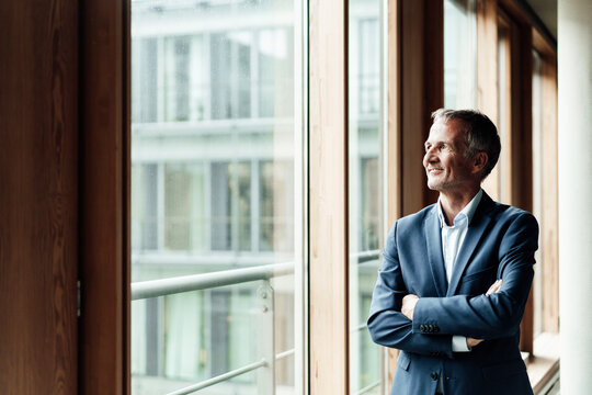 Contemplating businessman looking away while standing with arms crossed at window in office